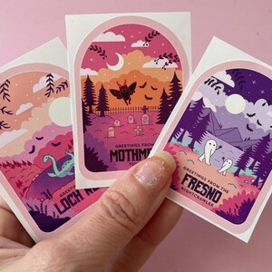 K&ouml;nnte beinhalten: Drei illustrierte Aufkleber mit einem rosa, violetten und orangenen Farbschema. Jeder Aufkleber zeigt eine andere gruselige Szene mit einem Monster, einem Geist und einer Kreatur aus dem Loch Ness. Die Aufkleber haben den Text "Greetings from the Mothman Town", "Greetings from the Fresno Nightcrawler" und "Greetings from Loch Ness".