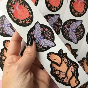 May include: A sheet of stickers featuring various designs. One sticker shows a human heart with floral accents. Another displays a moth with purple wings and a floral background. A third sticker depicts a hand holding a snake, surrounded by flowers.