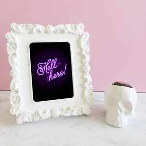 Puede incluir: Un marco de fotos blanco adornado con una foto en blanco y negro en su interior. La foto muestra un letrero de ne&oacute;n que dice "Hello there!" en morado. El marco est&aacute; sobre una superficie blanca junto a un cr&aacute;neo de cer&aacute;mica blanca con una peque&ntilde;a planta verde que crece en la parte superior.