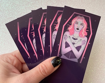 May include: A set of six bookmarks featuring a pink-haired woman in a coffin. The bookmarks are printed on a dark purple background with a white border.