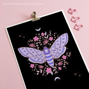 May include: A black background with a purple and blue moth with a skull on its head surrounded by pink and white flowers and gold leaves. There are gold crescent moons and stars in the corners.