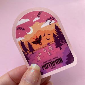 K&ouml;nnte beinhalten: Ein rosa und lilafarbener Aufkleber mit einer Mothman-Illustration und dem Text "Greetings from Mothman Town". Der Aufkleber zeigt einen Nachthimmel mit einer Mondsichel, Sternen und einem Friedhof mit einem Kreuz und Grabsteinen.