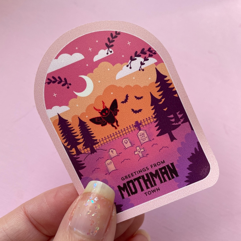 K&ouml;nnte beinhalten: Ein rosa und lilafarbener Aufkleber mit einer Mothman-Illustration und dem Text "Greetings from Mothman Town". Der Aufkleber zeigt einen Nachthimmel mit einer Mondsichel, Sternen und einem Friedhof mit einem Kreuz und Grabsteinen.