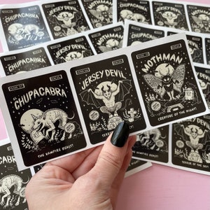 May include: A set of black and white stickers featuring illustrations of mythical creatures. The stickers are labeled with the names of the creatures, including "El Chupacabra", "Jersey Devil", and "Mothman".
