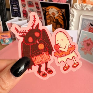 May include: A die-cut sticker featuring a dark brown moth creature with red eyes and antennae wearing orange shorts and sandals while holding an ice cream cone, next to a cream-coloured ghost wearing a red polka-dot floatie and sandals.