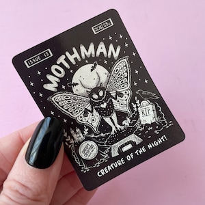 May include: Black and white illustration of a mothman creature with large wings, a heart on its chest, and the text "MOTHMAN CREATURE OF THE NIGHT!"  The illustration is on a black card with the text "ISSUE 13" and "CREEPY CRYPTIDS" in the bottom left corner.