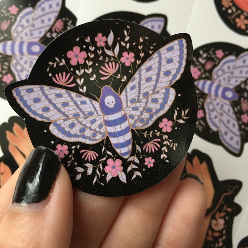 May include: Round black sticker featuring a moth with a skull design on its head and light purple wings. The moth is surrounded by pink flowers and leaves. The sticker is held in a hand.