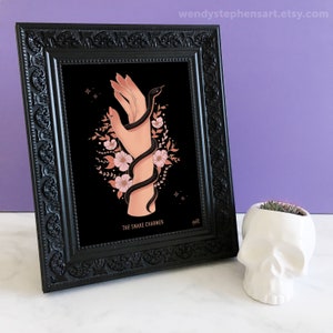 May include: A black framed print with a black snake wrapped around a hand with pink flowers. The print has the text "The Snake Charmer" at the bottom.