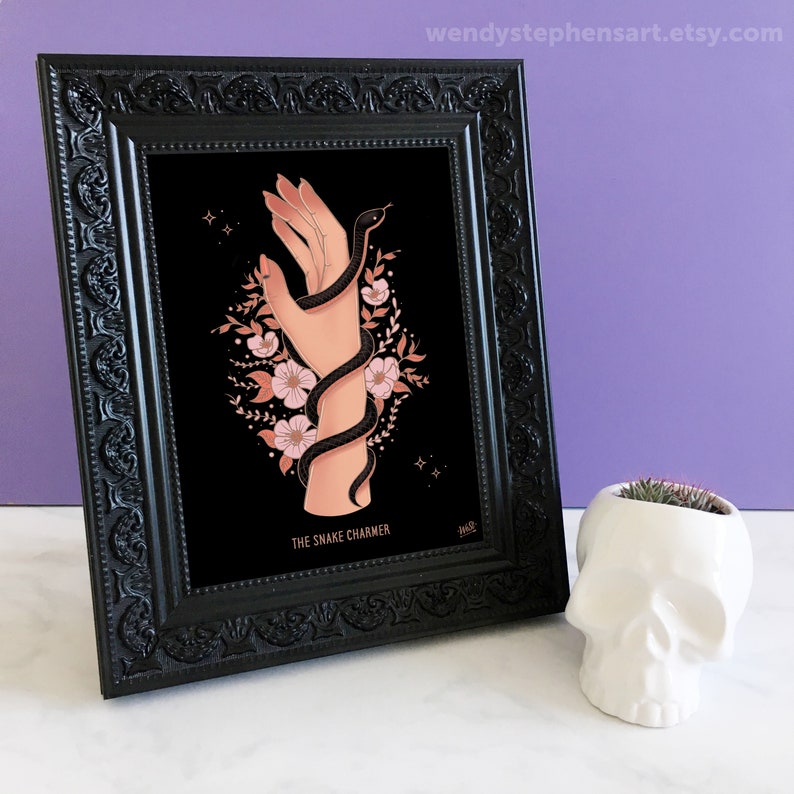 May include: A black framed print with a black snake wrapped around a hand with pink flowers. The print has the text "The Snake Charmer" at the bottom.