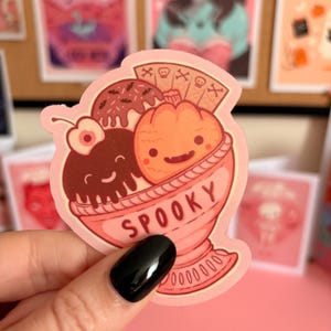 May include: A die-cut sticker featuring a pink sundae bowl labelled SPOOKY, containing a smiling chocolate ice cream scoop with a cherry, a smiling orange pumpkin scoop, and a wafer cookie decorated with skull and crossbones symbols.