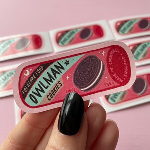 May include: A pink and blue sticker with the text "Owlman: Folklore Food Cookies" and a black and white illustration of an owl cookie. The sticker also includes the text "Made in Cornwall, England, Since 2009".