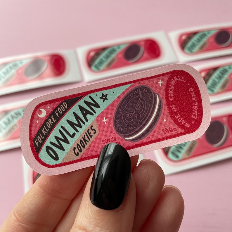May include: A pink and blue sticker with the text "Owlman: Folklore Food Cookies" and a black and white illustration of an owl cookie. The sticker also includes the text "Made in Cornwall, England, Since 2009".