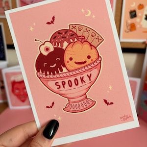 May include: A rectangular greeting card with a pink background featuring a cartoon illustration of a sundae in a bowl labelled SPOOKY. The sundae includes a chocolate scoop with a cherry, a pumpkin scoop, and a wafer cookie with skull icons.