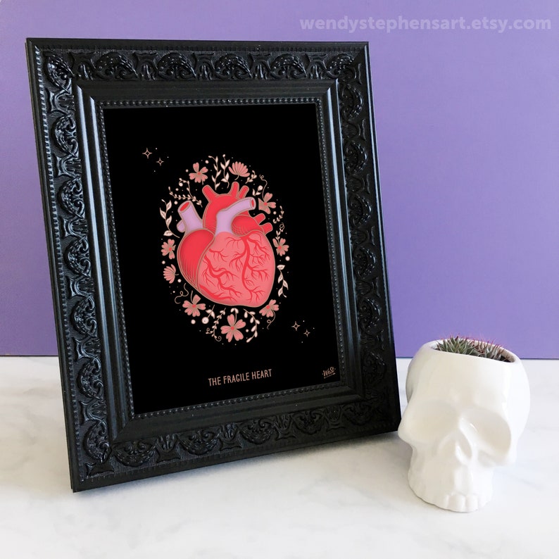 May include: A framed print with a black background featuring a red anatomical heart surrounded by pink and gold floral details. The text "The Fracile Heart" is printed below the image.