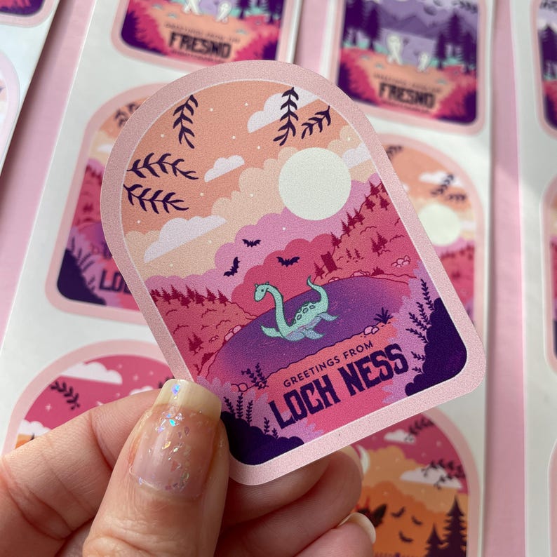 K&ouml;nnte beinhalten: Ein rosa und lilafarbener Aufkleber mit einer Cartoon-Illustration eines gr&uuml;nen Seeungeheuers aus Loch Ness in einem See. Der Aufkleber hat einen wei&szlig;en Mond und Flederm&auml;use am Himmel. Der Text "Greetings from Loch Ness" ist auf dem Aufkleber gedruckt.