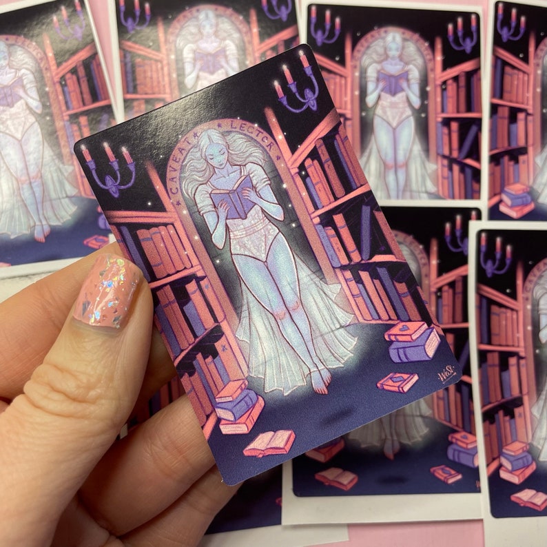 Puede incluir: Una pegatina con una figura fantasmal vestida con un vestido blanco leyendo un libro en un entorno de biblioteca. La figura est&aacute; rodeada de estanter&iacute;as llenas de libros. La pegatina tiene un esquema de color rosa y morado y el texto "Caveat Lector" est&aacute; escrito en un c&iacute;rculo alrededor de la figura.