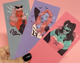 May include: Three illustrated bookmarks featuring stylized female figures. One is red-skinned with devil horns, another is blue-skinned with the text "UNLUCK 13", and the third is green-skinned with stitches. Each has a unique design and color scheme.