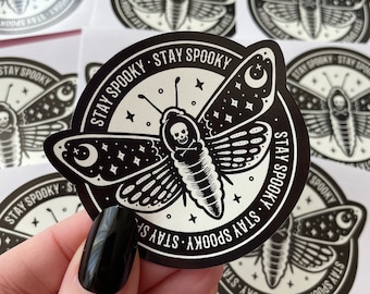 May include: A black and white sticker with a moth design. The moth has a skull and crossbones on its body and is surrounded by stars and a crescent moon. The text "Stay Spooky" is written around the moth in a circle.