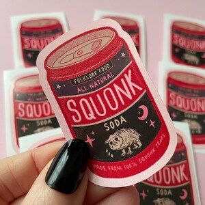 May include: A pink and red can of soda with the words "Folklore Food All Natural Squonk Soda" and a cartoon creature with a teardrop on its cheek. The can is surrounded by stars and a crescent moon.