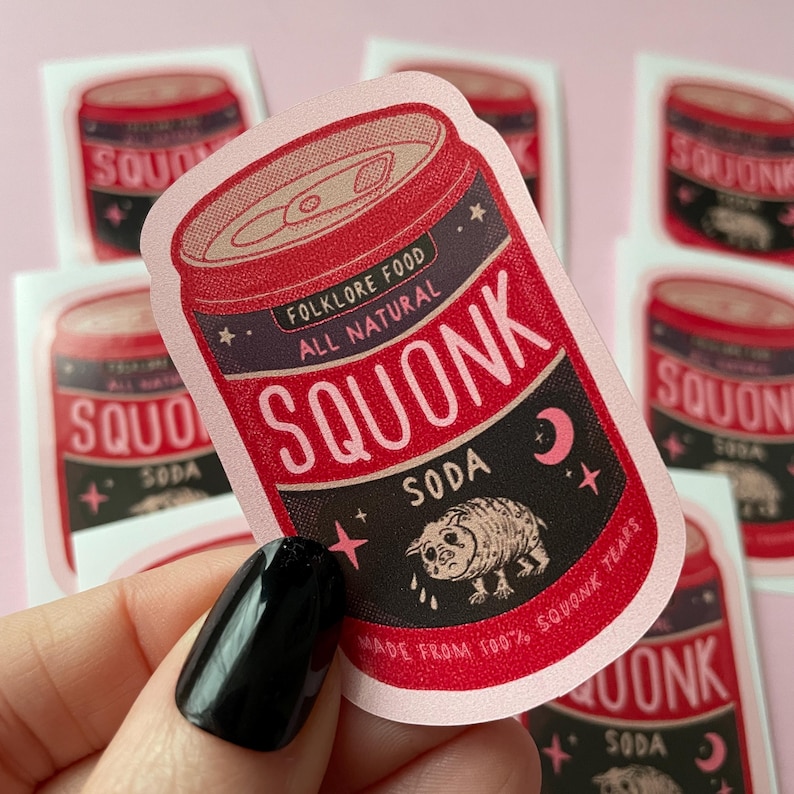 May include: A pink and red can of soda with the words "Folklore Food All Natural Squonk Soda" and a cartoon creature with a teardrop on its cheek. The can is surrounded by stars and a crescent moon.