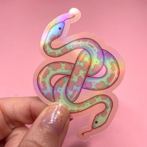 May include: Holographic sticker of a cartoon snake with a light green body and pink heart-shaped markings. The snake has a blue head and a red tongue. The sticker has a rainbow sheen and is on a pink background.
