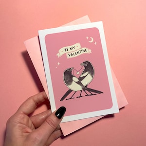 May include: A pink Valentine's Day card with a white border featuring two black and white birds holding a heart. The card has the text "BE MY VALENTINE" on a banner. A pink envelope is behind the card.