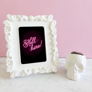 Puede incluir: Marco de fotos blanco adornado con una foto en blanco y negro en su interior. La foto muestra texto de ne&oacute;n rosa que dice "Hello there!" sobre un fondo de madera oscura. Un macetero de calavera blanco con una peque&ntilde;a planta verde est&aacute; sentado sobre una superficie blanca.
