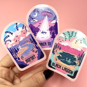 May include: Three illustrated stickers with a retro, vintage aesthetic. Each sticker features a different landscape with a mythical creature. The first sticker is pink and purple and says "Greetings from Bigfoot Country". The second sticker is blue and purple and says "Greetings from Area 51". The third sticker is green and pink and says "Greetings from the Black Lagoon".