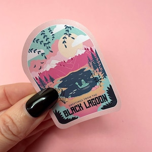 K&ouml;nnte beinhalten: Ein rosa und blauer Aufkleber mit einem Monster aus dem Cartoon in einem See, umgeben von Bergen. Der Aufkleber lautet "Greetings from the Black Lagoon".