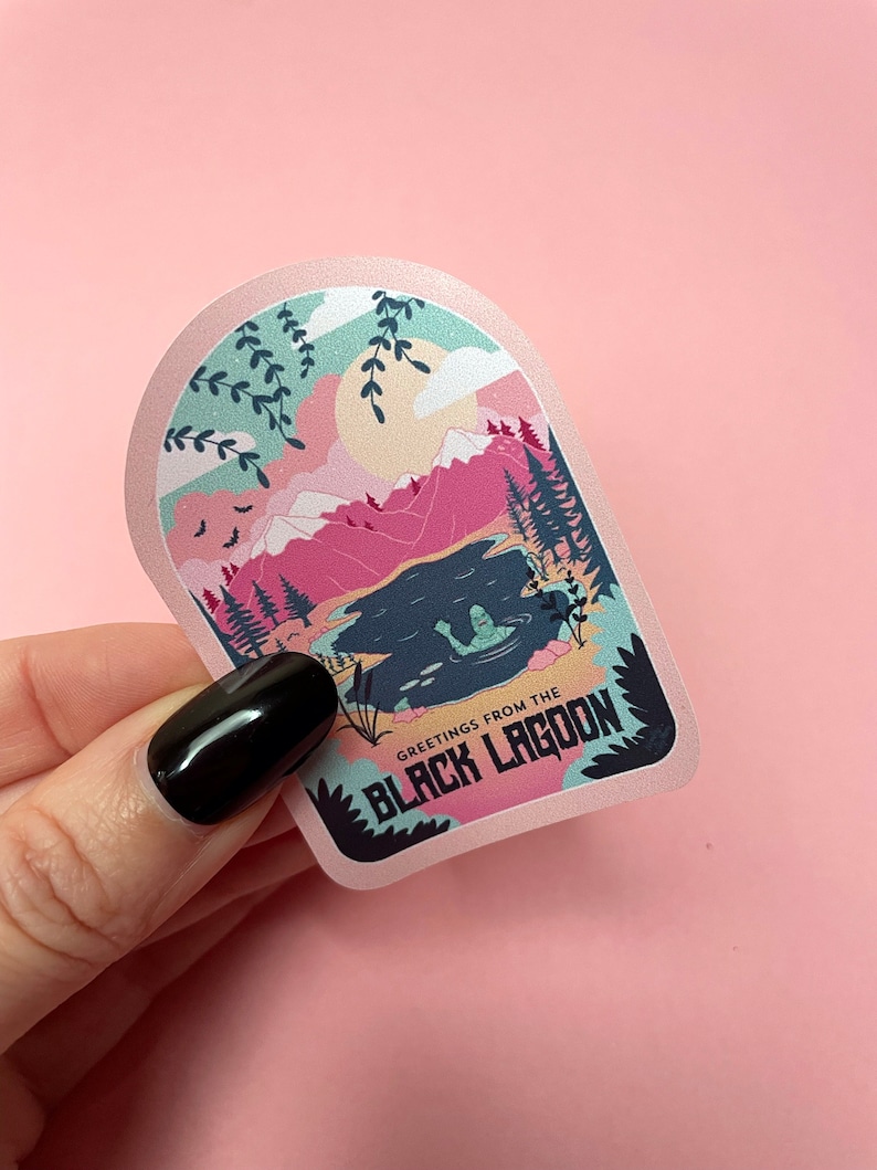 K&ouml;nnte beinhalten: Ein rosa und blauer Aufkleber mit einem Monster aus dem Cartoon in einem See, umgeben von Bergen. Der Aufkleber lautet "Greetings from the Black Lagoon".