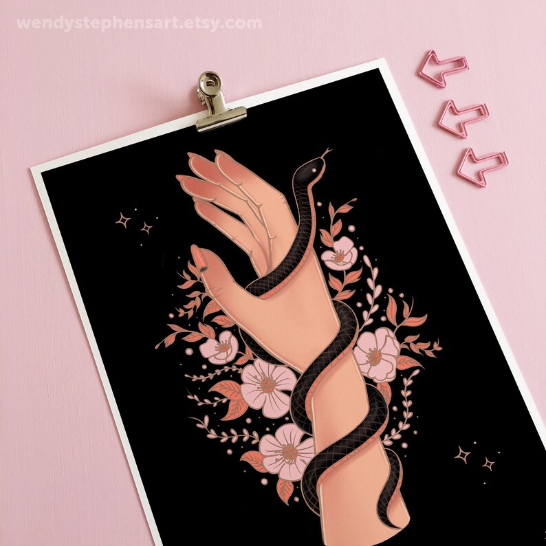 May include: A black snake wrapped around a hand with pink flowers and leaves on a black background. The snake has a red stripe down its back. There are two gold stars in the top left corner.