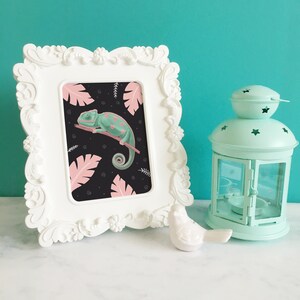 May include: A white ornate frame with a print of a green chameleon on a black background with pink tropical leaves. The chameleon has pink spots and is curled up on a branch.