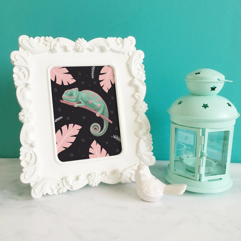 May include: A white ornate frame with a print of a green chameleon on a black background with pink tropical leaves. The chameleon has pink spots and is curled up on a branch.