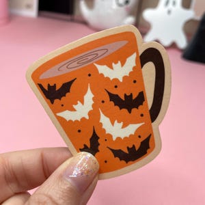 May include: A Halloween-themed sticker in the shape of a coffee mug. The mug is orange with white and black bat silhouettes. The mug handle is brown. The sticker is held by a hand with glittery nail polish.