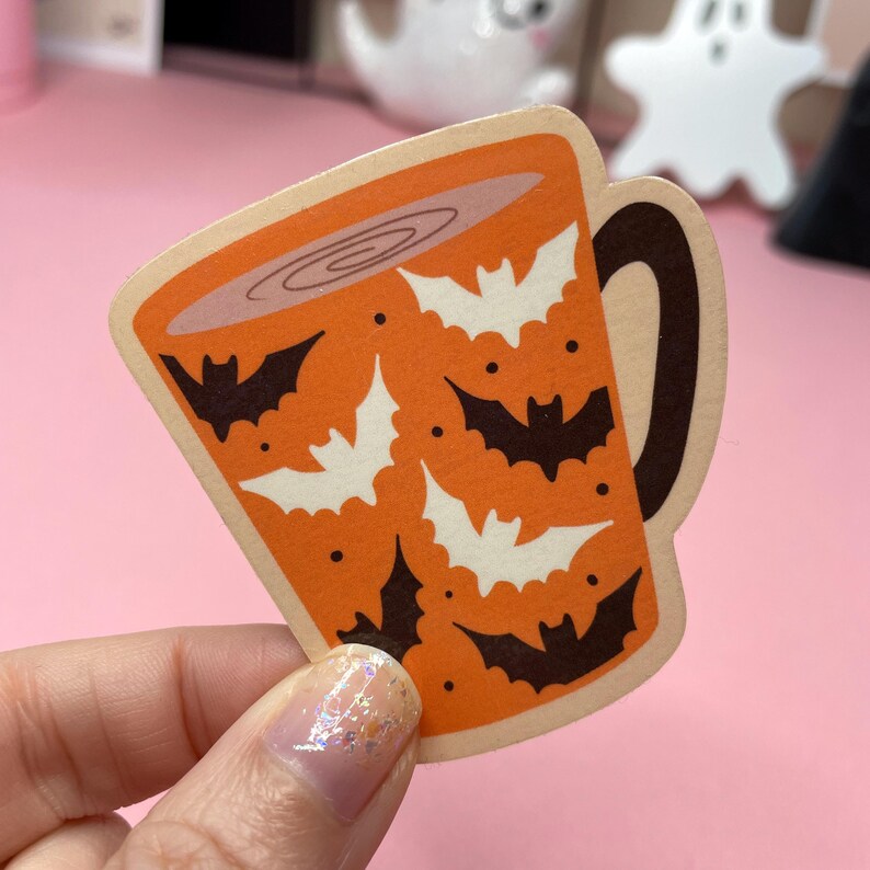 May include: A Halloween-themed sticker in the shape of a coffee mug. The mug is orange with white and black bat silhouettes. The mug handle is brown. The sticker is held by a hand with glittery nail polish.