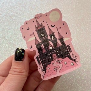 May include: A pink and black sticker featuring a spooky gothic castle with a clock tower, bats, and a ghost. The castle is surrounded by a graveyard with tombstones and a path leading to the front door. The sticker has a whimsical and cute aesthetic.