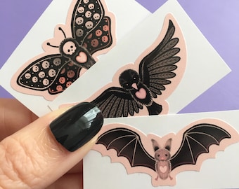 May include: Three stickers featuring black and pink illustrations of a butterfly, a bird, and a bat. Each illustration has a small pink heart on it.