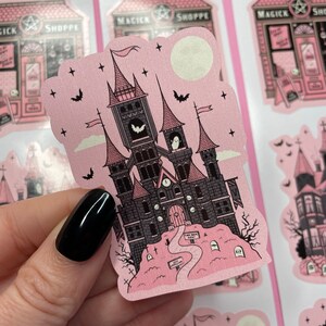 May include: Pink and black sticker featuring a spooky castle with a clock tower, bats, and a ghost. The castle is surrounded by a graveyard with tombstones and a path leading to the front door. The sticker has a glittery finish.