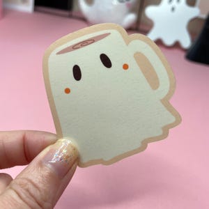 May include: A die-cut sticker in the shape of a coffee mug with a ghost-like design. The sticker is off-white with a light brown outline and a coffee-colored top. It features two black eyes and two orange cheek dots.