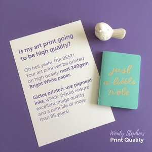 May include: A white card with text about high quality art prints. The text says "Is my art print going to be high quality? Oh hell yeah! The BEST! Your art print will be printed on high quality matt 240gsm Bright White paper. Giclee printers use pigment inks, which should ensure excellent image quality and a print life of more than 85 years!" A small teal notebook with gold lettering says "just a little note" and a white bird figurine is in the top right corner.