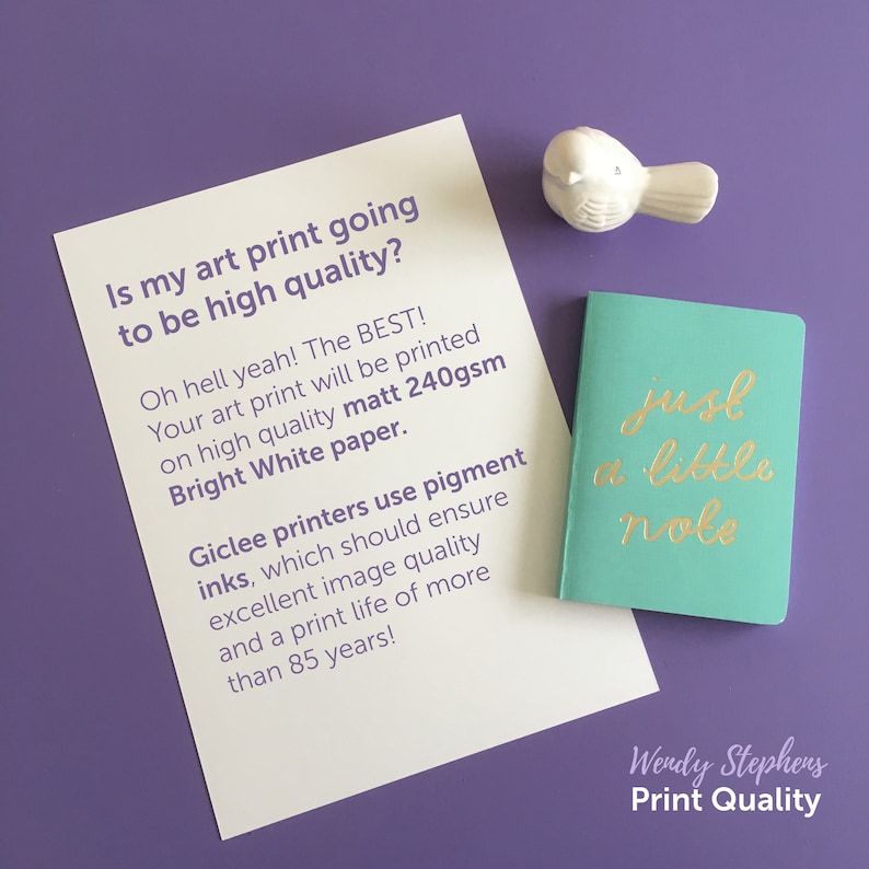 May include: A white card with text about high quality art prints. The text says "Is my art print going to be high quality? Oh hell yeah! The BEST! Your art print will be printed on high quality matt 240gsm Bright White paper. Giclee printers use pigment inks, which should ensure excellent image quality and a print life of more than 85 years!" A small teal notebook with gold lettering says "just a little note" and a white bird figurine is in the top right corner.