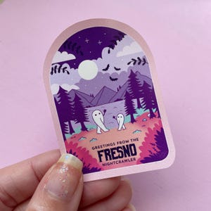K&ouml;nnte beinhalten: Ein rosa und lilafarbener Aufkleber mit einer Nachthimmel-Szene und zwei wei&szlig;en Geistern. Der Aufkleber lautet "Greetings from the Fresno Nightcrawler".