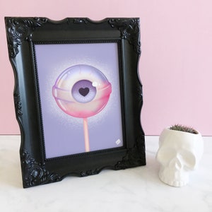 May include: A framed print of a colorful illustration of a lollipop with an eyeball and a heart in the center. The lollipop is on a stick and the background is a light purple color.