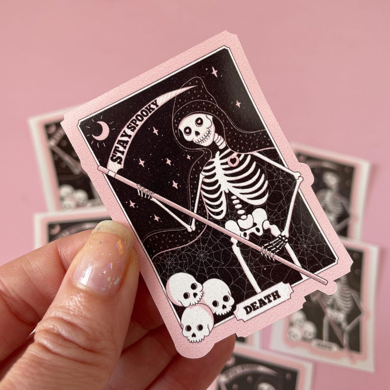 May include: Pink and black sticker with a skeleton holding a scythe. The sticker has the text "Stay Spooky" and "Death" on it.