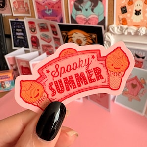 May include: A die-cut sticker with a light pink background featuring the red text Spooky Summer inside a decorative banner. Two orange soft-serve ice cream cones with smiling faces and the word Spooky printed on the cones flank the text.