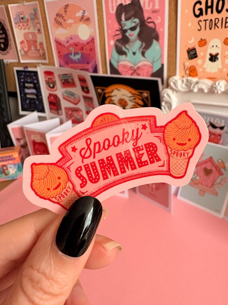May include: A die-cut sticker with a light pink background featuring the red text Spooky Summer inside a decorative banner. Two orange soft-serve ice cream cones with smiling faces and the word Spooky printed on the cones flank the text.