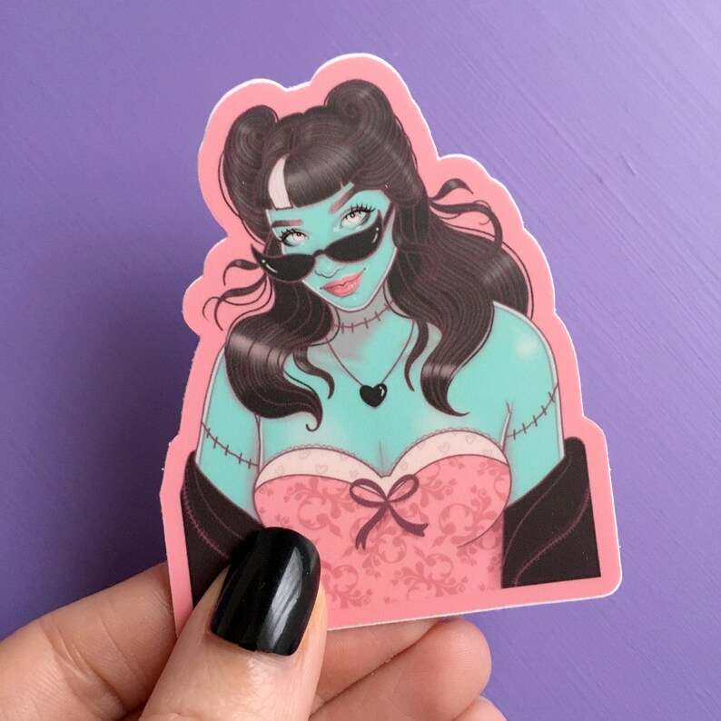 K&ouml;nnte beinhalten: Ein Aufkleber mit einer stilisierten Illustration einer Frau mit t&uuml;rkisfarbener Haut, schwarzem Haar und Sonnenbrille. Sie tr&auml;gt ein rosa Kleid mit Schleife und eine schwarze Herz-Halskette. Der Aufkleber hat einen rosa Rand.
