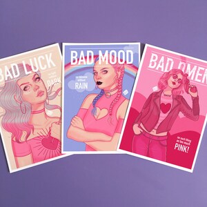 Puede incluir: Tres tarjetas coloridas con las palabras "Bad Luck", "Bad Mood" y "Bad Omen" impresas en ellas. Cada tarjeta presenta una ilustraci&oacute;n diferente de una mujer con cabello rosado y un atuendo rosado. El texto en las tarjetas incluye frases como "no light without dark", "no rainbow without rain", y "no such thing as too much pink!"