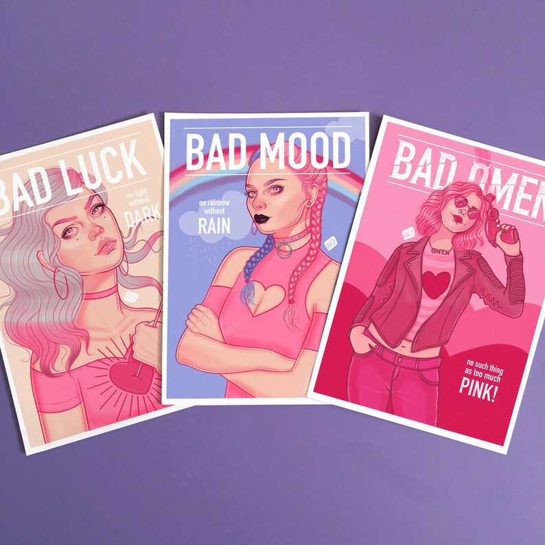 Puede incluir: Tres tarjetas coloridas con las palabras "Bad Luck", "Bad Mood" y "Bad Omen" impresas en ellas. Cada tarjeta presenta una ilustraci&oacute;n diferente de una mujer con cabello rosado y un atuendo rosado. El texto en las tarjetas incluye frases como "no light without dark", "no rainbow without rain", y "no such thing as too much pink!"