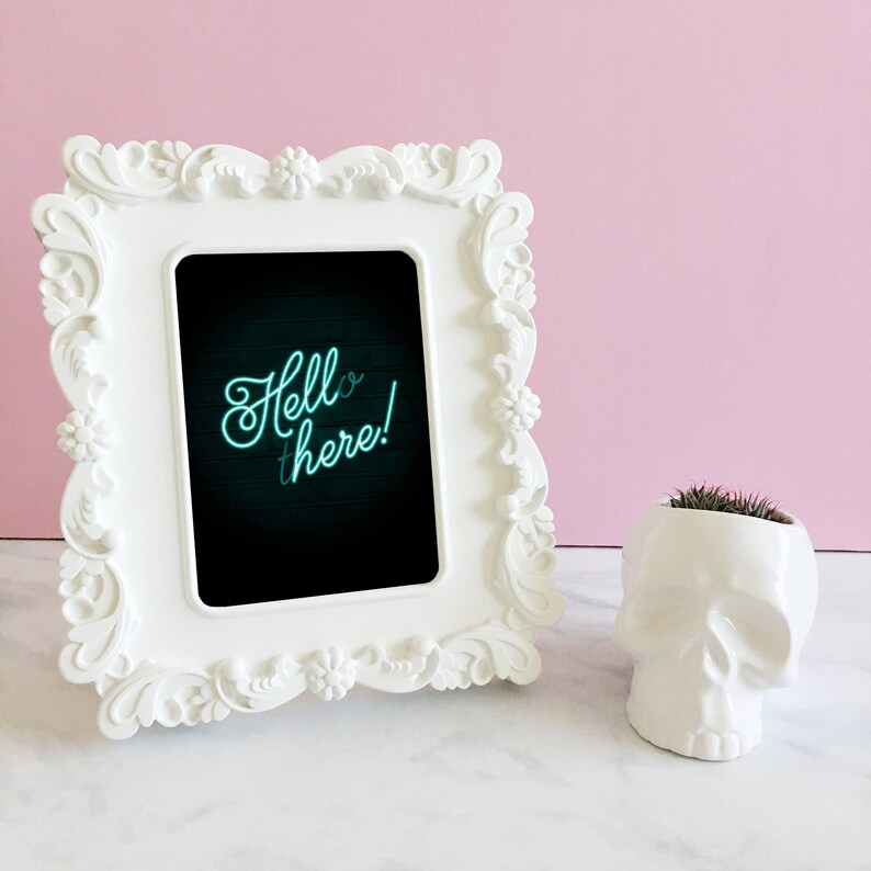 Puede incluir: Marco de fotos blanco adornado con una foto en blanco y negro en su interior. La foto dice "Hello there!" en una fuente verde ne&oacute;n. Un macetero de calavera blanco con una peque&ntilde;a planta verde est&aacute; en primer plano.
